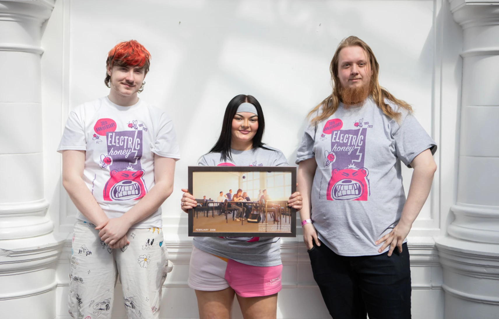 Three students wearing Electric Honey t-shirts standing in front of a white wall, holding a framed classroom photo from 2008. Three students wearing Electric Honey t-shirts standing in front of a white wall, holding a framed classroom photo from 2008.